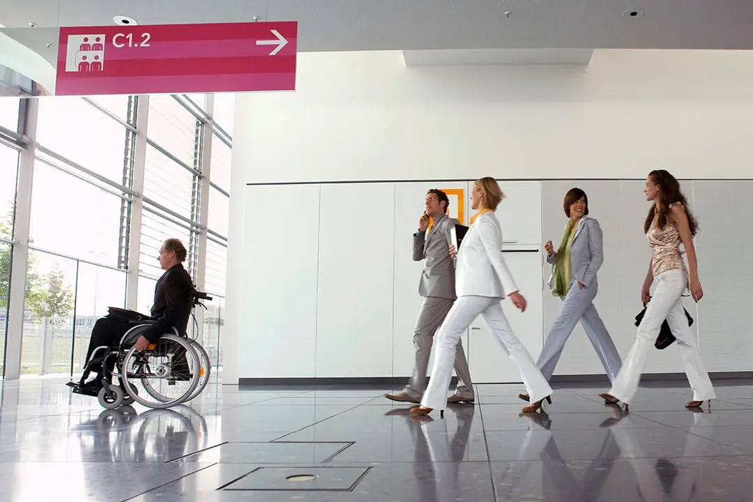 Placas de señalización direccional en el pasillo de una clínica moderna, indicando la ubicación de consultas, aseos y salidas de emergencia.