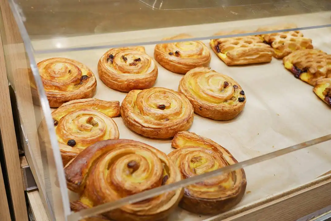 Vitrina de metacrilato para alimentos exhibiendo dulces y bollería en el mostrador de una cafetería moderna.